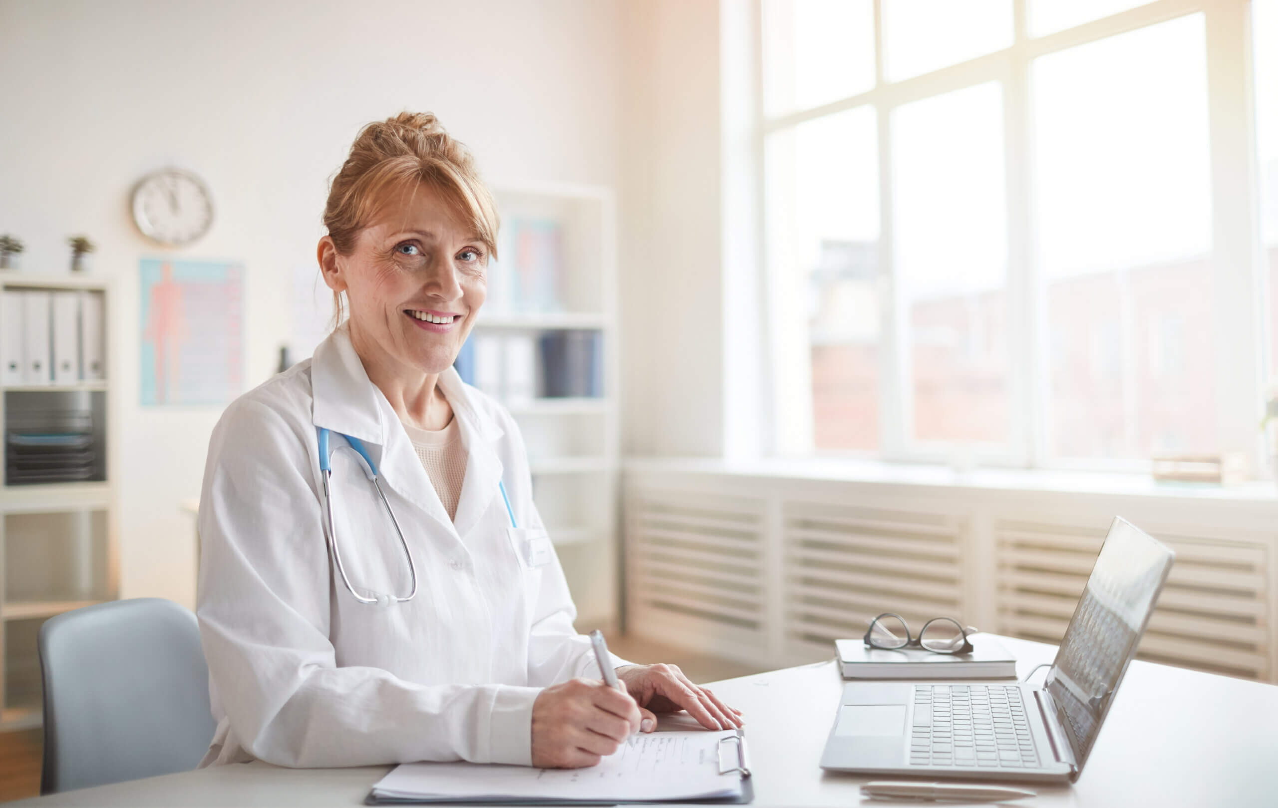 Portrait of mature female doctor in white coat smiling at camera while working at her workplace with documents